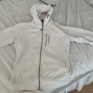Vit fleecejacka från Sail racing  - Vit fleecejacka från Sail racing med hög krage och justerbar huva. Koftan har hel dragkedja framtill och en bröstficka med dragkedja. Perfekt för kyliga dagar och sportiga outfits.