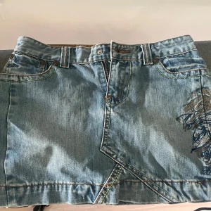 Broderad jeanskjol från Miss Sixty - Säljer en kort jeanskjol från Miss Sixty med snygg broderad detalj i lila, guld, blått och rött på sidan. Kjolen har klassisk femficksdesign, bälteshällor och dragkedja framtill. .