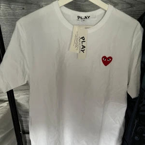 Vit t-shirt Comme des Garçons Play - Vit t-shirt från Comme des Garçons Play med klassisk röd hjärtlogga med ögon broderad på bröstet. T-shirten har rund hals och korta ärmar, tillverkad i mjuk bomull. Perfekt för dig som gillar stilrena och ikoniska plagg. Aldrig använd då den är för stor.