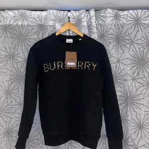 Svart Burberry sweatshirt med logga - Svart sweatshirt från Burberry med klassisk rund hals och lång ärm. Stor Burberry-logga i rutigt mönster framtill. Tröjan har ribbade muddar vid ärmslut och nederkant. Perfekt för dig som gillar stilrena designer med lyxig touch.