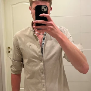 River Island beige långärmad skjorta  - Stilren beige skjorta från River Island med klassisk krage och knappar hela vägen. Liten broderad logga på bröstet och diskret randig detalj på insidan av knappslån. Skjortan är i mjuk bomull och har lång ärm med knapp vid manschetten.