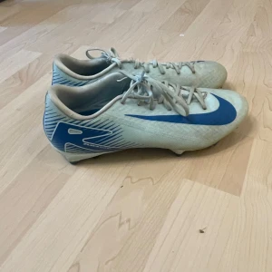 Nike fotbollsskor i vitt och blått utan skål låda - Snygga fotbollsskor från Nike i vitt med blå detaljer och stor blå swoosh på sidan. Skorna har snörning och dobbar för bra grepp på planen. Lätta och smidiga med sportig design, perfekta för dig som vill sticka ut på matchen.