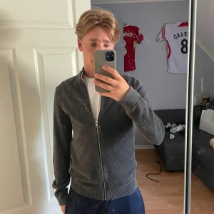 Grå zip-kofta från Jack & Jones - Snygg grå kofta från Jack & Jones med hel dragkedja och ribbad krage. Modellen är långärmad och har en stilren, enkel design som passar till många olika outfits. Materialet är stickat och känns mjukt mot huden. Perfekt för dig som gillar klassisk stil.