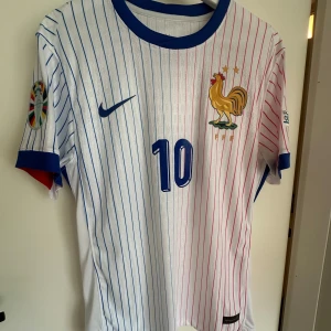 Kylian Mbappe Frankrike EM 2024 Autentisk Kvalité  - Snygg vit fotbollströja från Nike med tunna blå och röda ränder, blå krage och ärmslut. Franska landslagets emblem med tupp och två stjärnor på bröstet, samt tryck med nummer 10 och Mbappé på ryggen. Materialet är lätt och ventilerande polyester, perfekt för match eller träning.