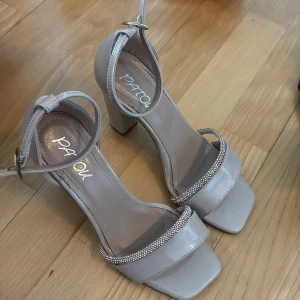 Beige sandaler med klack från Patou - Använt 1 gång. Stilrena beige sandaler från Patou med bred rem över tårna dekorerad med glittrande stenar. Skorna har fyrkantig tå, hög blockklack och justerbar ankelrem med spänne. Tillverkade i glansigt syntetmaterial som ger en lyxig känsla.