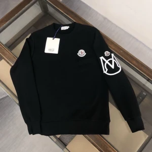 Svart sweatshirt från Moncler - Svart sweatshirt från Moncler med klassisk rund hals och långa ärmar. På bröstet och ena ärmen sitter Moncler-loggor, samt en stor vit M-design på ärmen. Tröjan har en clean look och är perfekt för dig som gillar stilrena märkesplagg.
