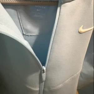 Nike NOCTA ljusblå zipjacka - Ljusblå zipjacka från Nike NOCTA med stilren design och diskret Nike-logga på bröstet. Jackan har hel dragkedja, hög krage och långa ärmar. Perfekt för dig som gillar sportig och clean stil.