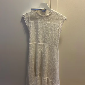 Vit spetsklänning med öppen rygg - Superfin vit klänning i spets med blommigt mönster och volangdetaljer vid ärmslut. Klänningen har en snygg öppen rygg och är ärmlös med rund halsringning. Perfekt för sommarens alla tillfällen!