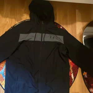 Svart vindjacka från Under Armour - Svart vindjacka från Under Armour med grått band och logga över bröstet. Jackan har huva med snörning, dragkedja framtill och långa ärmar. Perfekt för dig som vill ha en sportig och clean look.