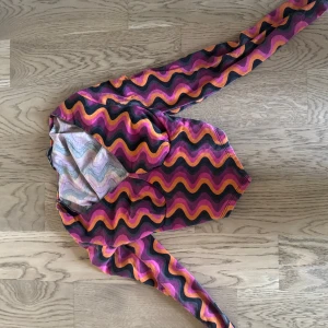 Färgglad långärmad topp - Säljer en långärmad topp i storlek XXS med vågigt mönster i lila, rosa, orange och svart. Toppen har en figurnära passform och är tillverkad i ett mjukt, stretchigt material som känns skönt mot huden. Perfekt för dig som gillar färg och retro vibes!