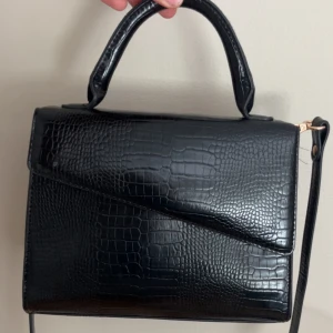 Svart handväska i croco-imitation - Stilren svart handväska med croco-mönstrad yta och elegant handtag. Väskan har en asymmetrisk lockdesign och avtagbar axelrem i samma material. Perfekt för dig som gillar en klassisk look med modern twist.