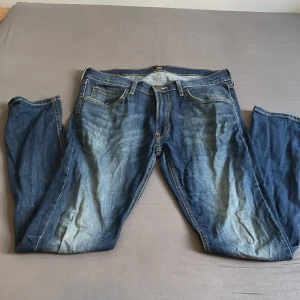 Blå Lee jeans med raka ben - Säljer ett par klassiska blå jeans från Lee med raka ben och normal passform. Jeansen har snyggt slitna detaljer framtill, fem fickor och Lee-logga på bakfickan. Perfekta för dig som gillar en tidlös denimlook.