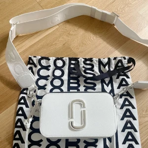 Vit axelväska från Marc Jacobs - LÅNADE BILDER från hon jag köpte från som köpte från mintoo. Snygg vit axelväska från Marc Jacobs med bred justerbar axelrem och silverfärgade detaljer. Väskan har dubbla dragkedjor och ikonisk logga framtill. Tillverkad i skinn och perfekt för att lyfta din outfit med en clean och modern look. Äktighets bevis finns på väskan!