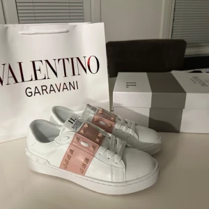 Valentino Rockstud sneakers vit/rosa - Snygga Valentino Rockstud sneakers i vitt skinn med ljusrosa band och ikoniska nitar. Klassisk rund tå, platt sula och vita snören. Perfekt för dig som vill ha en lyxig och trendig look med en touch av rosa detaljer.