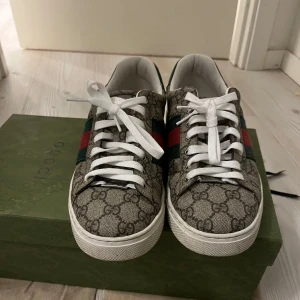 Gucci sneakers med GG-mönster - Snygga Gucci sneakers med klassiskt GG-monogram i beige och brunt, gröna och röda ränder på sidorna och vita snören. Skorna har rund tå, platt sula och är tillverkade i canvas med skinn-detaljer. Perfekta för dig som vill sticka ut med lyxig streetstyle.Ny pris 7000