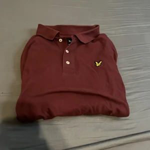 Vinröd pikétröja från Lyle & Scott - Snygg vinröd pikétröja från Lyle & Scott med klassisk krage och tre knappar framtill. Tröjan har det ikoniska gula fågel-loggan broderad på bröstet. Perfekt för dig som vill ha en stilren och bekväm look. Hör av er vid minsta lilla fråga 