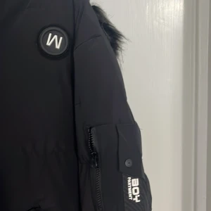 Svart pufferjacka med fuskpäls från BOY - Svart pufferjacka från BOY med huva och snygg fuskpälsdetalj. Jackan har dragkedja, flera fickor med coola detaljer och BOY-märke på ärmen. Perfekt för kalla dagar och riktigt street i stilen.