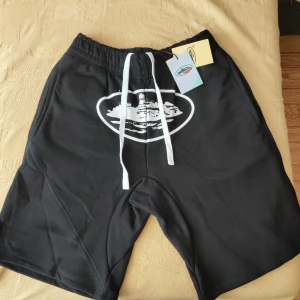 Svarta Cortiez Shorts (S) - Svarta shorts från Cortiez med vitt grafiskt tryck framtill och breda vita snören i midjan. Modellen har elastisk midja och en bakficka med lock. Materialet är mjukt och skönt, perfekt för chill dagar. Snygg streetwear vibe och riktigt bekväma.