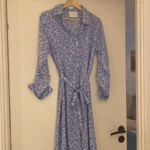 Blå blommig långklänning Sisters Point - Supersöt blå långklänning från Sisters Point med vitt blommigt mönster. Klänningen har långa ärmar, krage, knappar framtill och ett matchande knytbälte i midjan. Perfekt för dig som gillar en feminin och fräsch stil.