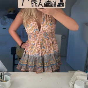 Blommig kortklänning från Lita Tomato - Supersöt kortklänning från Lita Tomato med blommigt mönster i blått, gult och beige på ljusrosa botten. Djup v-ringning, volangkant nertill och öppen rygg med knyt i nacken. Perfekt för sommarens varma dagar!