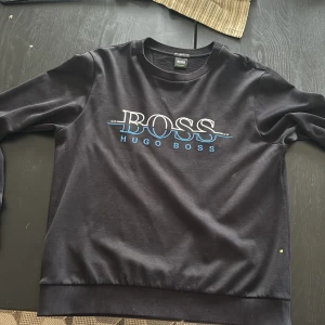 Svart Hugo Boss sweatshirt slim fit - Svart sweatshirt från Hugo Boss med stort BOSS-tryck i vitt och blått framtill. Slim fit-modell med rund hals och långa ärmar. Tillverkad i mjuk bomull för en skön känsla. Perfekt för dig som gillar stilren och clean streetwear.