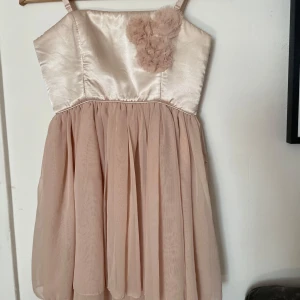 Champagnefärgad kort klänning med blommor - Supersöt kort klänning i champagnefärg med glansig överdel och fluffiga blommor framtill. Kjolen är i flera lager och axelbanden är smala. Köpt på bikbok för länge sedan men bara använd 1 gång🩷🤍