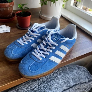 Adidas Gazelle blå mockasneakers - Adidas Gazelle sneakers i blå mocka med vita detaljer och klassiska tre ränder på sidorna. Skorna har vit hälkappa, vit snörning och gummisula med mönster. Perfekt för dig som gillar retrostil och vill sticka ut med färg. Skorna är använda lite så mockan är väldigt lite skadad.