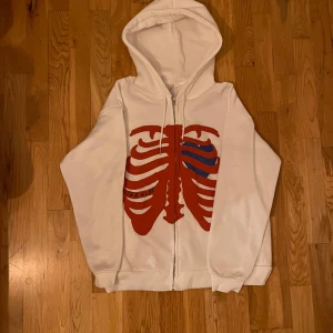 ÄKTA Vit Ver Blee hoodie (ORG PRIS 800 KR) - Säljer en vit hoodie med dragkedja och ett unikt tryck av röda och blå revben på framsidan. Perfekt för en cool och avslappnad stil. Hoodien har en justerbar huva och långa ärmar.