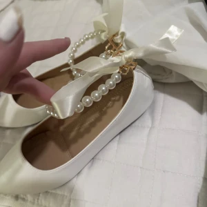 Vita ballerinaskor med pärlband och rosett - Supersöta vita ballerinaskor med spetsig tå, dekorerade med pärlband och satinrosett runt vristen. Skorna har en platt sula och är tillverkade i syntetmaterial med mjuk innersula för extra komfort. Perfekta för dig som vill ha en elegant och feminin look.