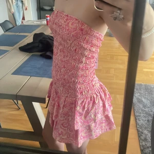 Rosa smockad tubklänning paisley S - Supersöt kort tubklänning med smockad överdel och volangig kjol. Klänningen har ett paisleymönster i rosa och ljusgult, är axelbandslös och sitter tajt upptill. Perfekt för sommardagar när du vill sticka ut lite extra! Säljs helt ny för den var för kort för mig