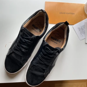 Svarta sneakers från Muzzo Footwear - Svarta sneakers från Muzzo Footwear med snygg mix av mocka och skinn. Klassisk design med rund tå, vita platta sulor och svarta snören. Perfekta för en clean och stilren look till vardags. Insidan är ljusbeige för extra kontrast.