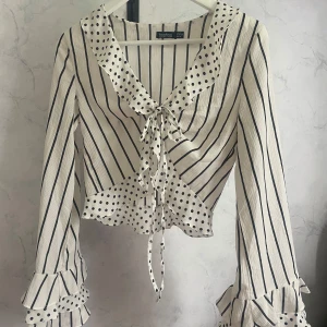 Randig och prickig blus från Boohoo - Supersöt blus i vitt med svarta ränder och prickar från Boohoo. Den har långa ärmar med volangdetaljer, knytning framtill och en mix av randigt och prickigt tyg på krage, ärmslut och nederkant. Croppad modell som ger en trendig look.