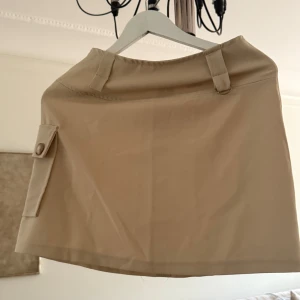 Beige kortkjol med ficka  - Stilren beige kortkjol med praktisk ficka på sidan med knappar och snygga bälteshällor. Kjolen har dragkedja i sidan och är gjord i ett slätt, lätt material som känns bekvämt. Perfekt för dig som gillar minimalistisk och clean stil. Ca 42 cm lång och ca 34 cm i midjan. 