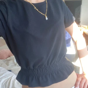 Svart tshirt smockad midja - Supersnygg svart blus med korta ärmar och smockad midja som ger en fin volangkant nedtill. Tunt, lätt material som känns luftigt och passar perfekt till jeans eller kjol. Enkel och stilren design som är lätt att matcha.