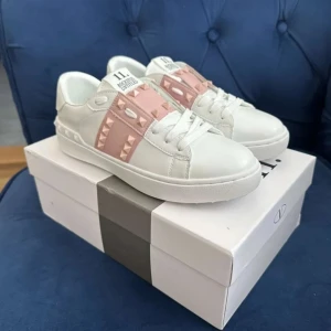 Valentino Rockstud sneakers vit/rosa - Snygga Valentino Rockstud sneakers i vitt skinn med ljusrosa detaljer och ikoniska nitar. Klassisk rund tå, platt sula och vita snören. Perfekta för dig som vill ha en lyxig och trendig look med en touch av rosa. Behöver sälja snabbt eftersom jag ska på semester om en vecka
