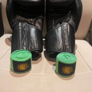 Svarta boxningshandskar Budo Nord - Säljer ett par svarta boxningshandskar från Budo Nord Fight Gear. Handskarna är tillverkade i skinn med perforerade detaljer och bred kardborrestängning. Medföljer gröna lindor med svart och gult lejonmärke. Perfekta för träning och sparring. Kan diskutera pris!