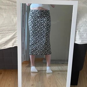 Svart blommig långkjol - Svart långkjol med vita små blommor i ett tätt mönster. Kjolen har en rak och figurnära passform som sitter snyggt i midjan och slutar vid vaden. Perfekt för dig som gillar en stilren men ändå söt look.