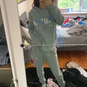 Grå trapstar hoddie med grå trapstar byxor  - Mysig grå hoodie med vit och blå text framtill. Tröjan har huva med snörning och en klassisk känguruficka. Perfekt för chill dagar och enkel att matcha med andra plagg. Långärmad och i mjukt material. Gråa mjukis byxor matchande med hoddie 