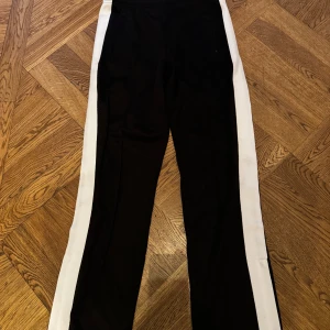 Svarta Sandro byxor - Svarta byxor från Sandro Paris med breda vita revärer längs sidorna. Snygg och sportig look med raka ben och elastisk midja. Perfekta för en trendig och avslappnad stil. Storlek S i mycket bra skick. 