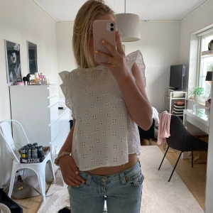 Vit blus med volang och broderi - Superfin vit blus med broderade blommor och volangdetaljer på axlarna. Blusen är ärmlös och har en söt liten krage. Perfekt till jeans för en fräsch look. Luftig och lätt i bomull, perfekt för varma dagar.