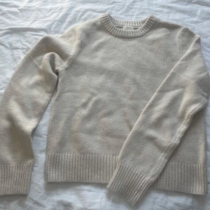 Beige stickad tröja med rund hals - En stilren beige stickad tröja med rund halsringning och breda ribbade muddar vid ärmslut och nederkant. Perfekt för lager-på-lager och enkel att matcha med jeans eller kjol. Mjuk och skön känsla, passar dig som gillar en clean och avslappnad look.