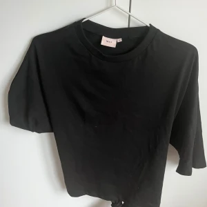 Svart knyt-topp från Nelly - Svart croppad topp från Nelly med korta ärmar och rund halsringning. Toppen har en snygg knyt-detalj i ryggen som ger en cool och trendig look. Perfekt att matcha med höga jeans eller kjol för en stilren outfit.