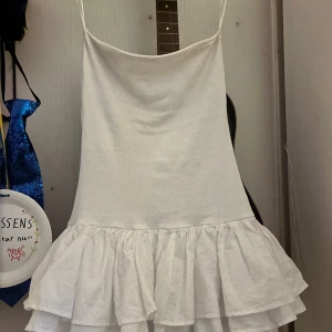 Vit kort volangklänning - Supersöt vit kort klänning med volangig kjol. Klänningen är figurnära upptill och har två lager volanger nertill som ger en fluffig och lekfull look. Perfekt för sommarens alla tillfällen och lätt att styla med smycken eller sneakers.