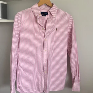 Rosa och vit randig skjorta Ralph Lauren - Säljer en klassisk rosa och vit randig skjorta från Ralph Lauren i modellen Custom Fit. Skjortan har lång ärm, knappar framtill och broderad logga på bröstet