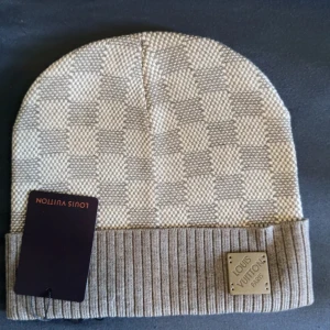 Beige rutig mössa  - Exklusiv beige och grå rutig mössa från   med klassiskt Damier-mönster. Mössan har ribbad uppvikt kant och en metallbricka med   Paris på framsidan. Perfekt accessoar för dig som vill sticka ut med lyxig stil.
