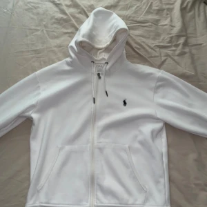 Vit hoodie från Polo Ralph Lauren - Vit hoodie från Polo Ralph Lauren med svart broderad logga på bröstet. Tröjan har dragkedja, snörning i huvan och två stora fickor framtill. Tillverkad i mjuk bomull, perfekt för chill dagar. Klassisk och stilren design. Dock är det en fläck där back men vet inte om den kan gå bort eller inte. 