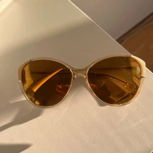 Guldiga solglasögon från Michael Kors - Snygga solglasögon från Michael Kors med transparent guldig båge och bruna linser. Cat-eye form med coola detaljer i guld på sidorna. Perfekta för att lyfta din stil och ge en lyxig känsla till din look.