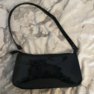 Svart axelväska i lack med dragkedja - Snygg svart axelväska i glansigt lackmaterial med minimalistisk design. Väskan har en smal axelrem, guldfärgade metalldetaljer och stängs med dragkedja upptill. Perfekt storlek för det viktigaste och ger en stilren vibe till din outfit.