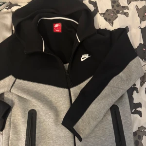 Nike svart och grå hoodiejacka M - Nike hoodiejacka i svart och ljusgrått med dragkedja hela vägen, huva och två fickor med dragkedja. Snygg sportig look med vit Nike-logga på bröstet. Tillverkad i mjuk polyesterblandning, perfekt för chill eller träning.
