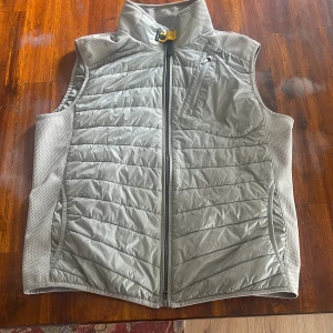Ljusgrå dunväst från Parajumpers XL - Snygg ljusgrå dunväst från Parajumpers i storlek XL. Västen har quiltad framsida och baksida, hög krage med gul detalj, dragkedja framtill och meshpaneler på sidorna. Perfekt lager-på-lager-plagg för kyliga dagar.